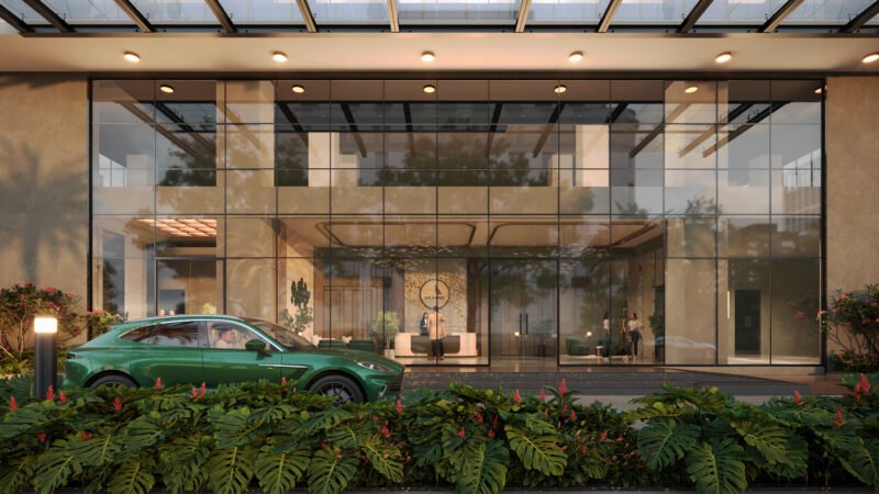 Front entrance of NRI Avenue building with a modern glass façade. A green car is parked in front, with lush landscaping and red flowers along the walkway. Inside, a reception area is visible through the glass, showing people interacting and a stylish, well-lit interior